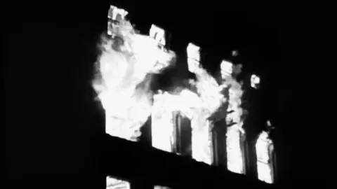 Black-and-white image showing a dark building interior with tall rectangular windows glowing bright white, while thick smoke or flames pour out through the openings, creating a stark, high-contrast scene that suggests a building on fire at night.